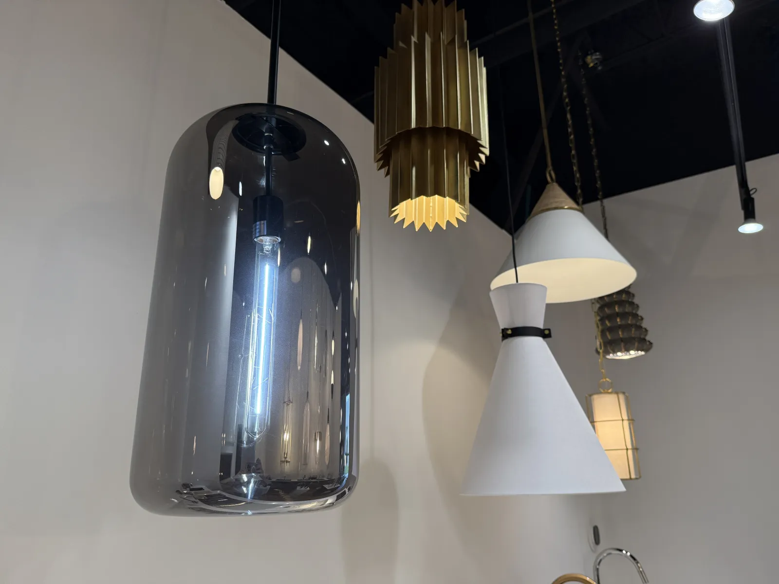 Modern hanging pendant lights with various shapes and materials in a showroom setting under ceiling spotlights.