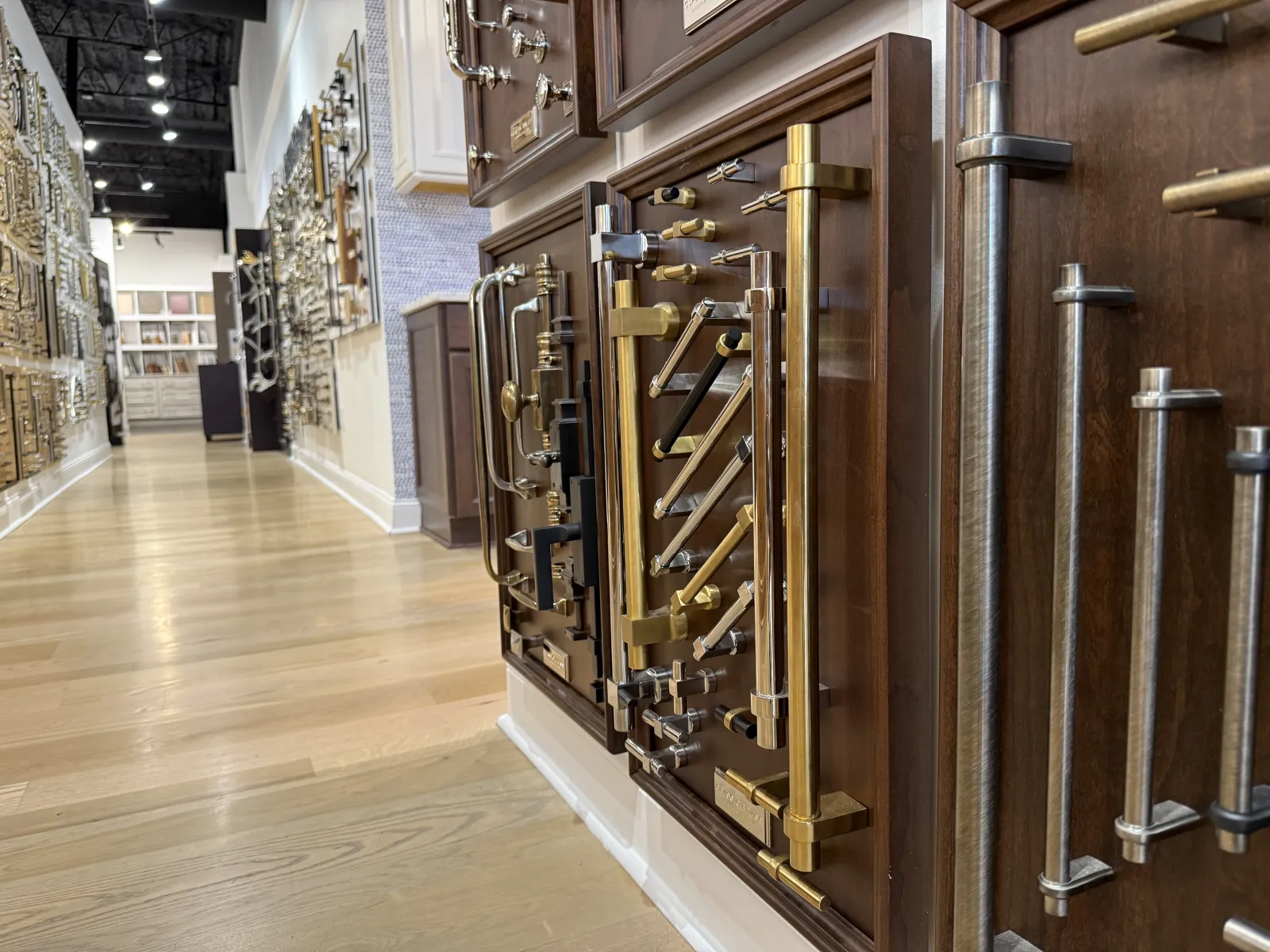Display of various metallic cabinet handles and pulls mounted on wooden panels in a showroom aisle.