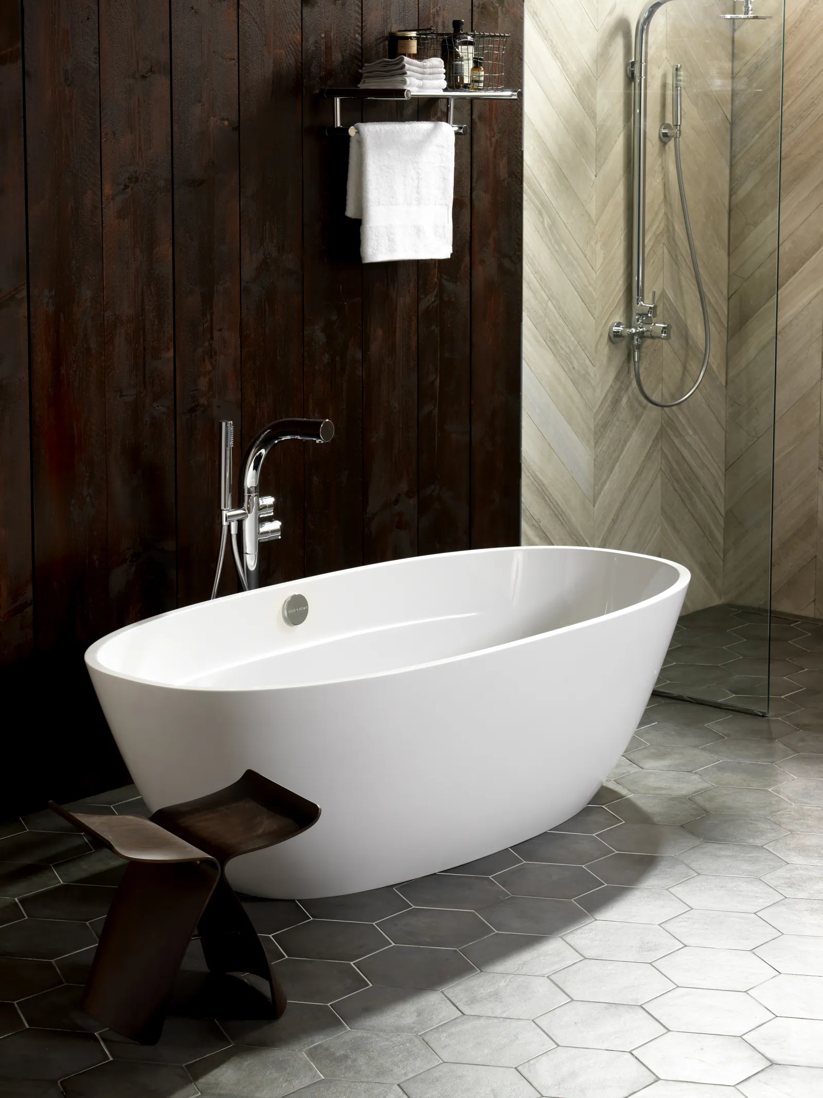 Modern bathroom with white freestanding bathtub, dark wood wall, hexagon floor tiles, and glass shower.
