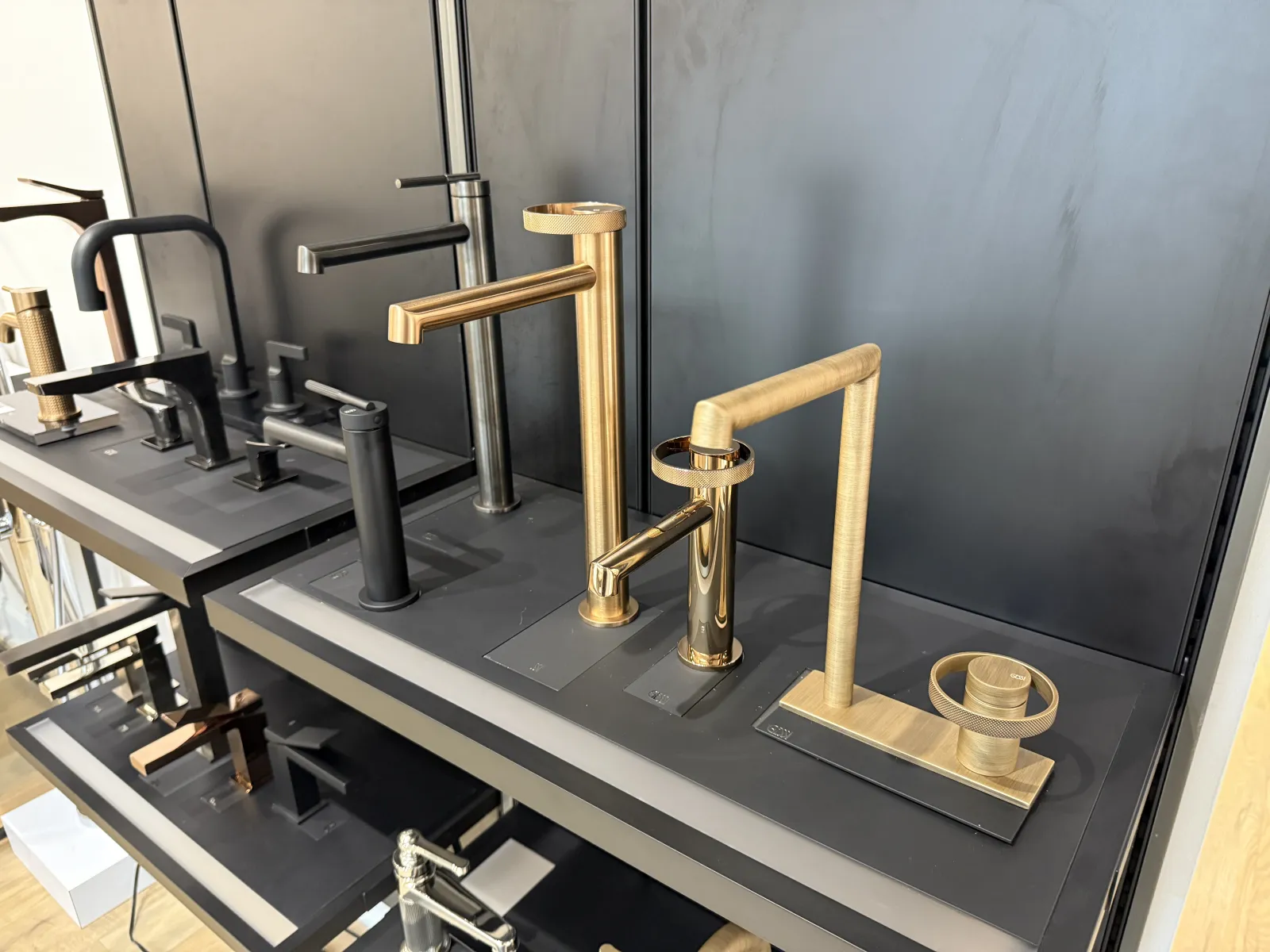 Modern matte black and gold kitchen faucets displayed on shelves in a showroom with a sleek black background.