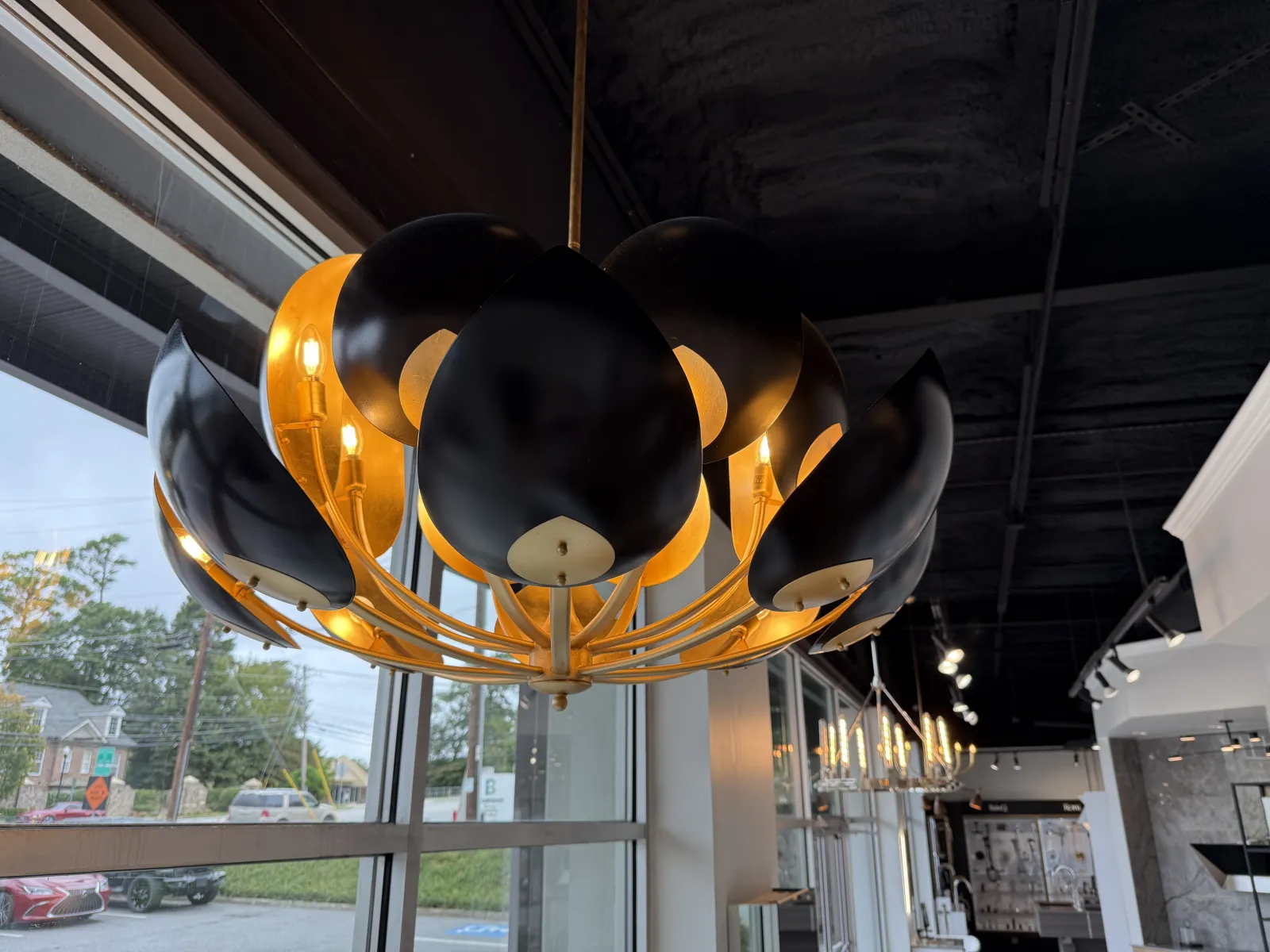 Modern black and gold chandelier with curved petals hanging near large windows in a showroom.