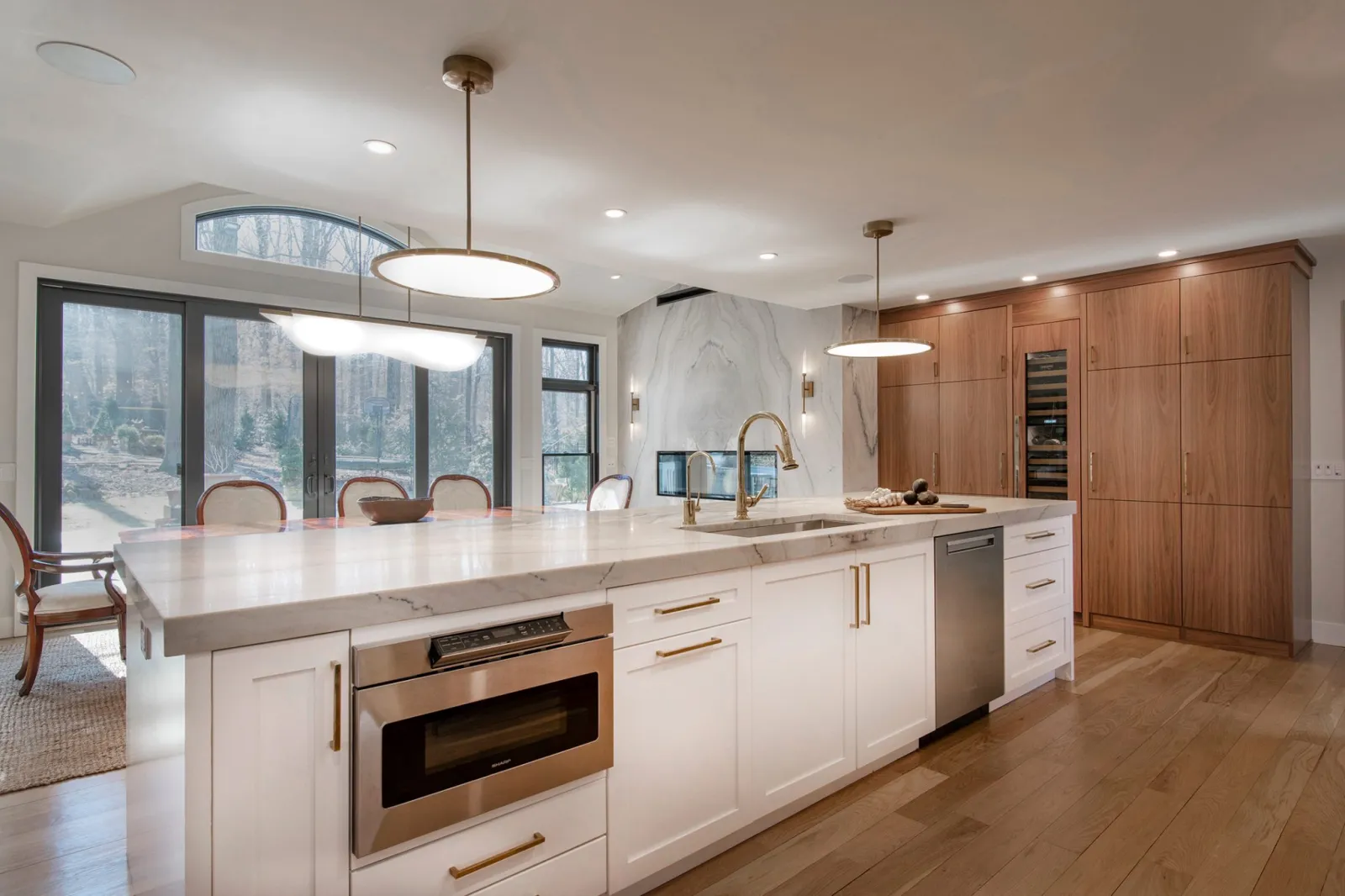 Modern kitchen with white marble island, brass fixtures, wood cabinets, stainless appliances, and large windows.