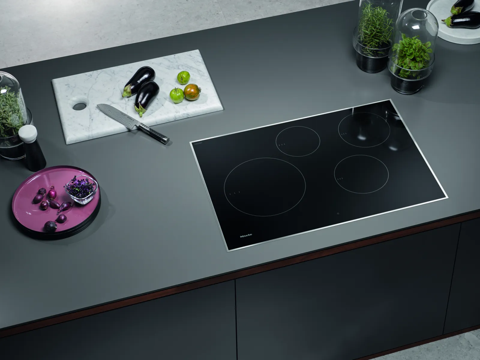 Modern kitchen with a sleek induction cooktop, vegetables, and a marble cutting board for food prep.