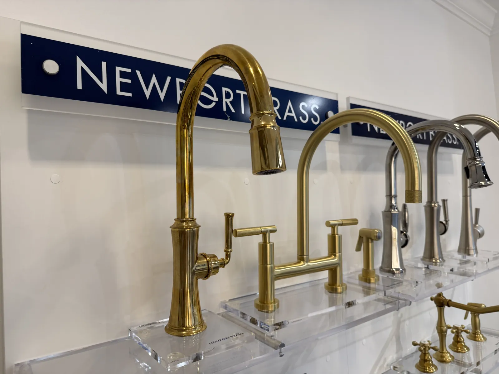 Display of brass and chrome kitchen faucets mounted on clear acrylic stands in a showroom.