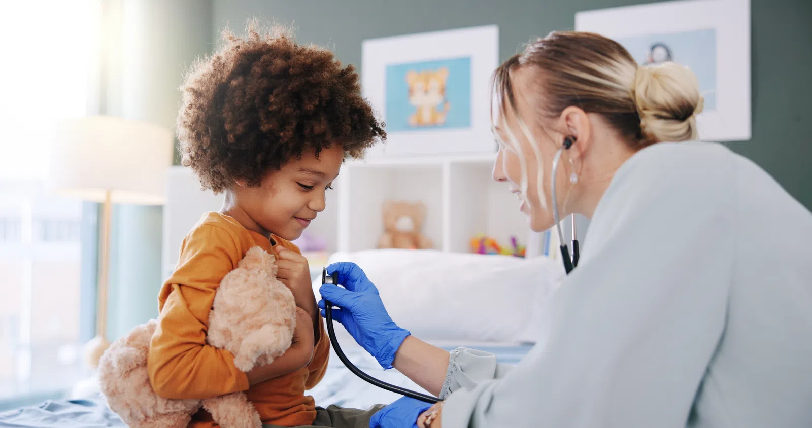 Doctor using stethoscope to check a smiling child's heartbeat while the child holds a teddy bear in a cozy room.