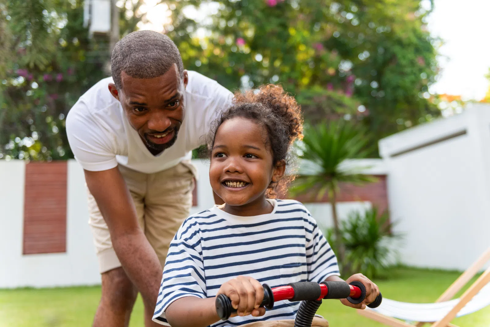Father supports smiling daughter riding a red tricycle in a sunny backyard with greenery and flowers.