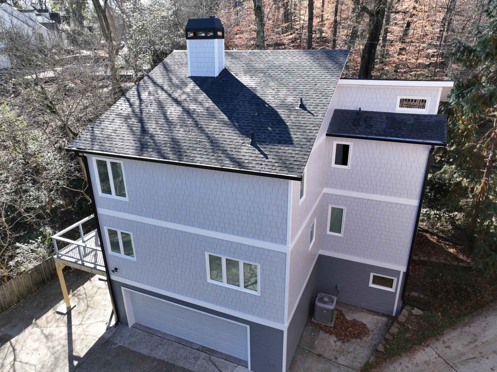 Modern three-story gray house with black roof, white trim, garage, and small balcony in wooded area.