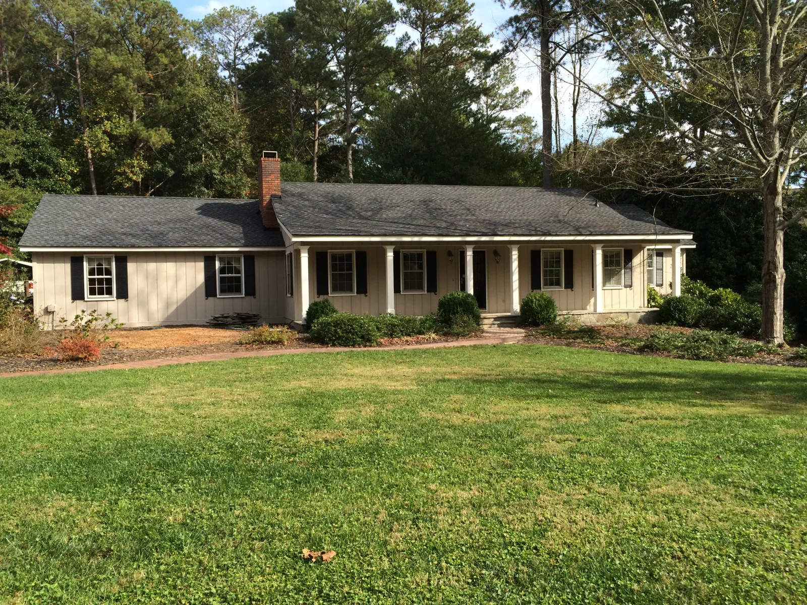Single-story beige house with black shutters, front porch, and large green lawn surrounded by tall trees.