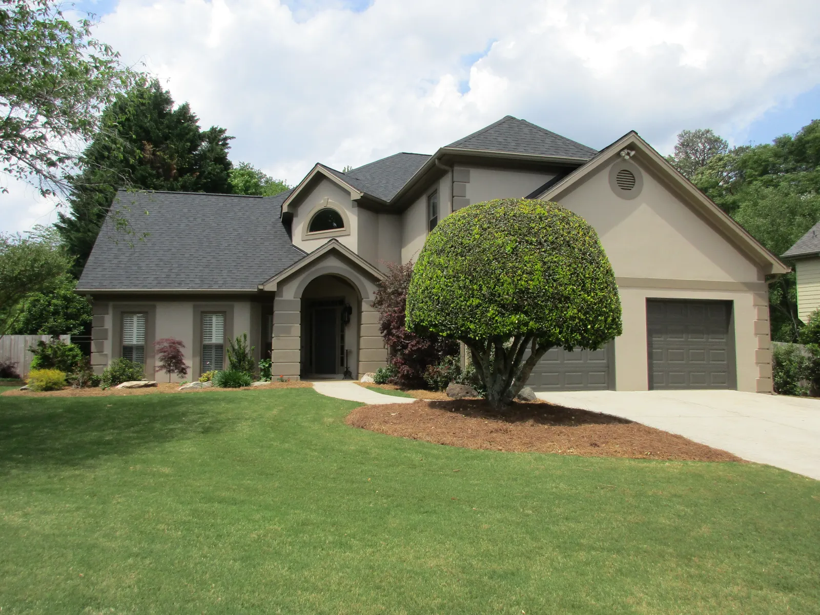 Modern beige two-story house with a large green lawn and uniquely trimmed round tree in front.