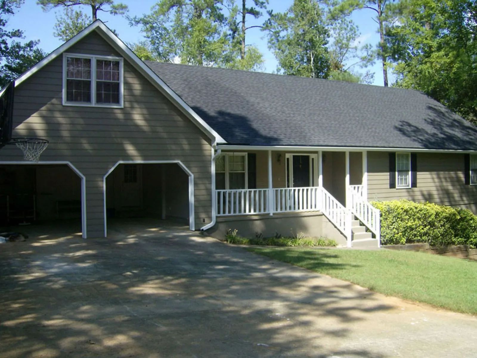 Single-story house with two-car garage, front porch with white railings, and surrounding trees on a sunny day