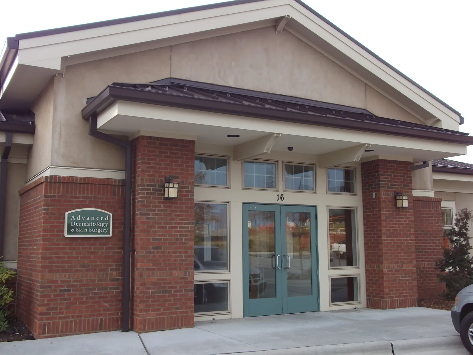 Entrance of Advanced Dermatology & Skin Surgery clinic with brick pillars and teal double doors under a gabled roof.