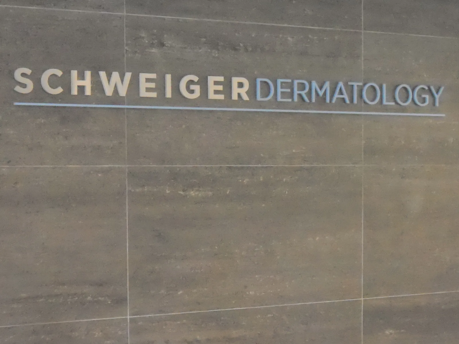 Schweiger Dermatology logo displayed on a gray tiled wall with beige and blue lettering.