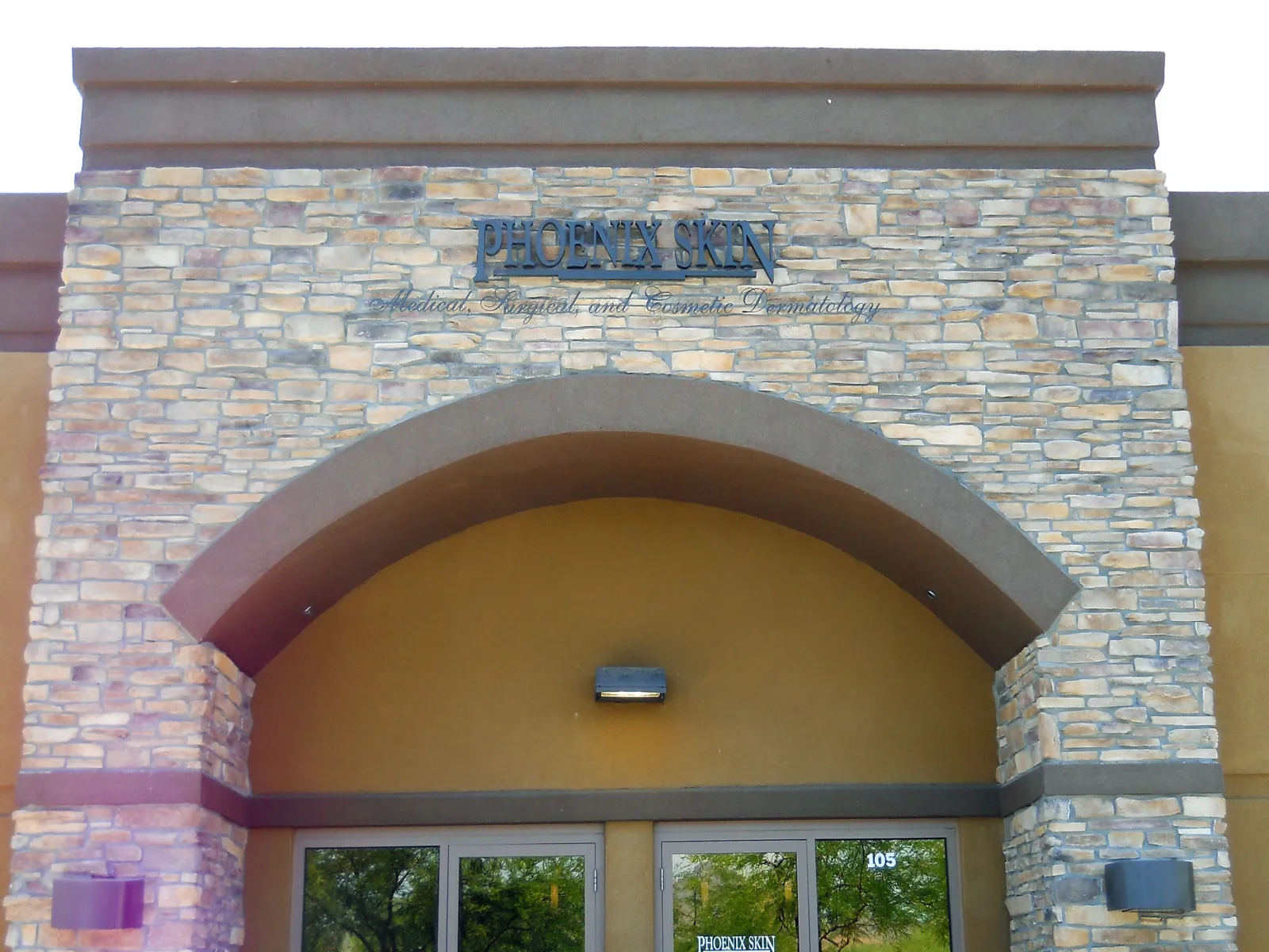 Stone facade and arched entrance of Phoenix Skin Dermatology clinic with glass doors reflecting trees.