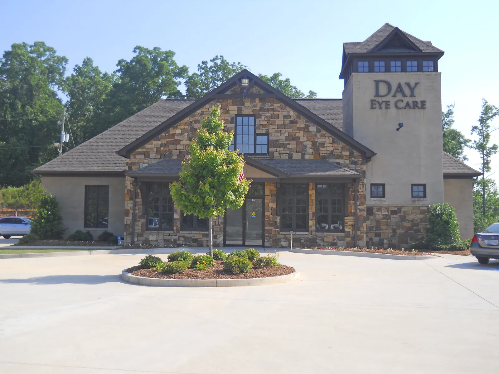 Modern stone and stucco eye care clinic building with a small tree and roundabout in front under clear sky