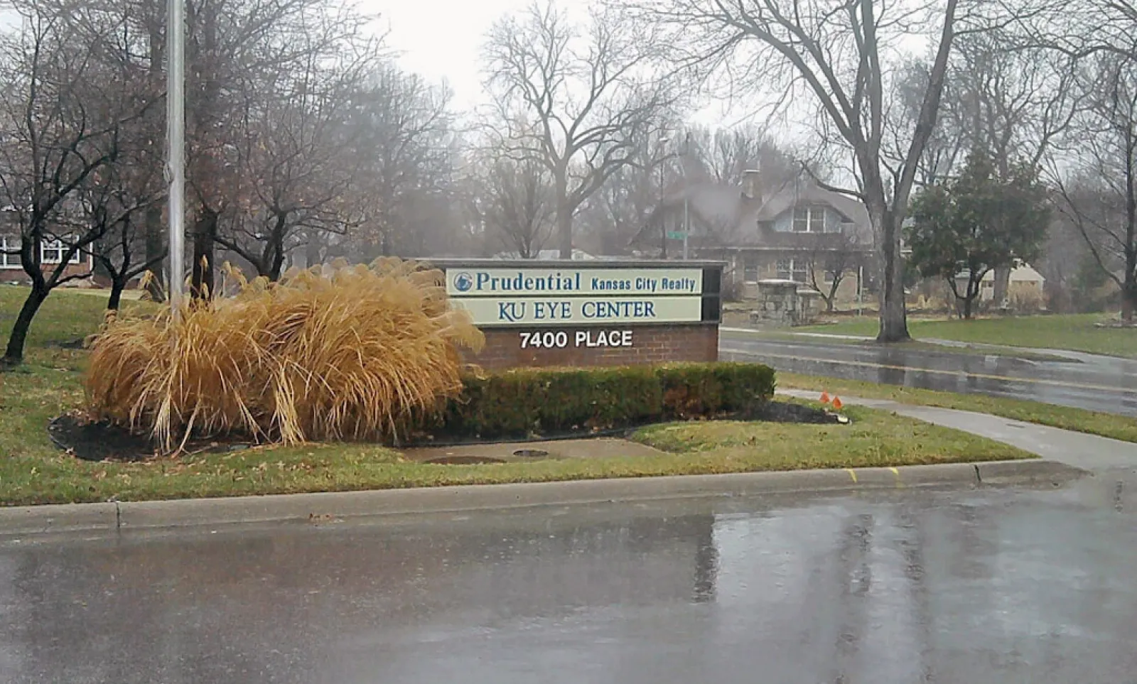 Sign for KU Eye Center and Prudential Kansas City Realty on a rainy day with bare trees and wet pavement.