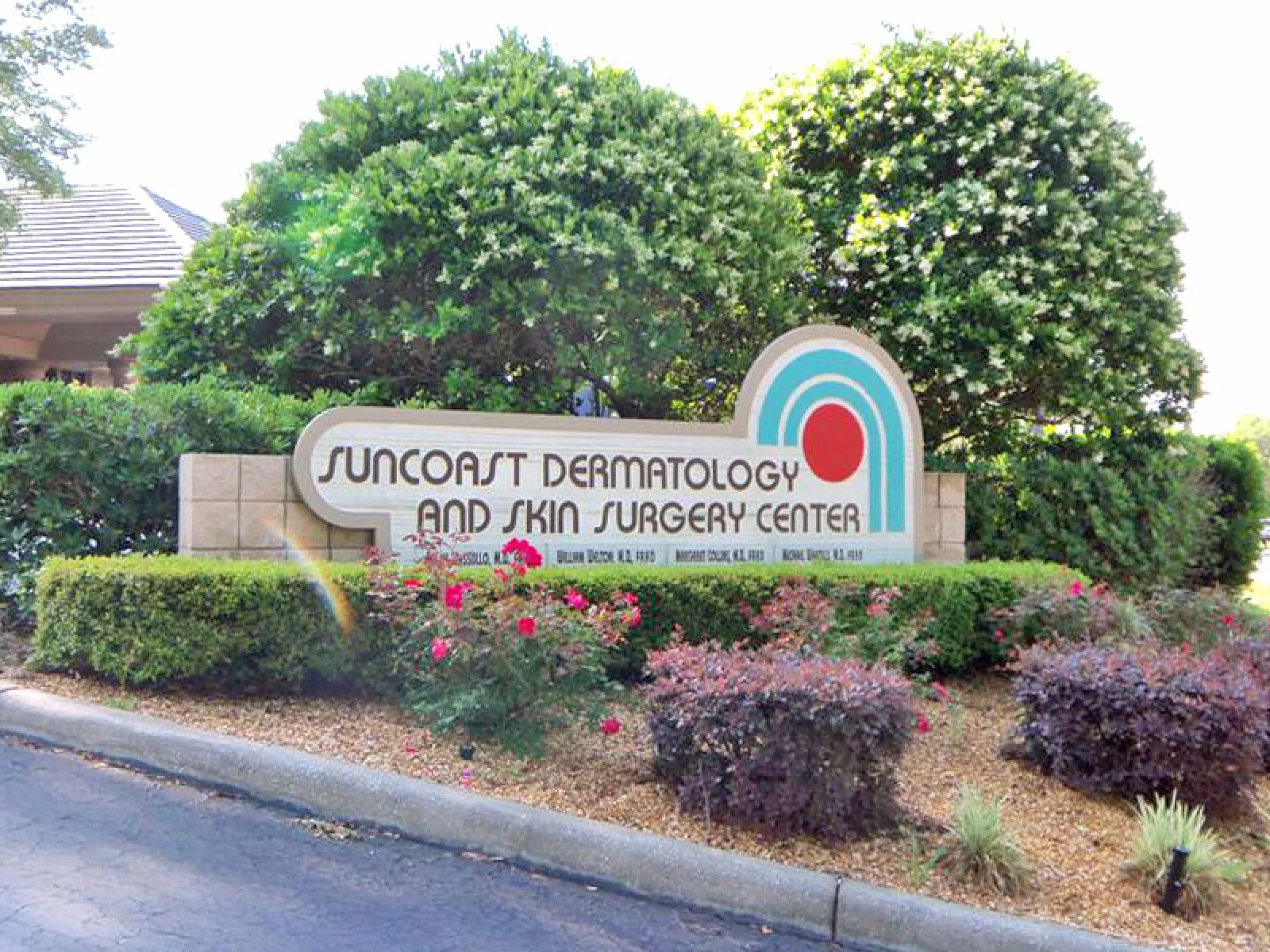 Sign for Suncoast Dermatology and Skin Surgery Center surrounded by bushes and flowers at roadside.