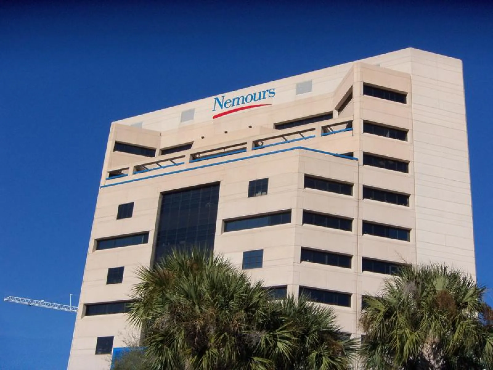 Modern beige office building with Nemours logo atop under clear blue sky and palm trees in foreground