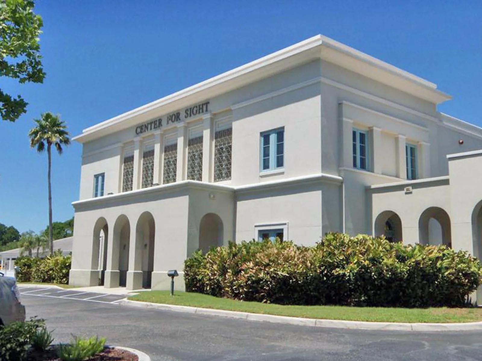White two-story building labeled Center for Sight with arched windows and greenery under clear blue sky.