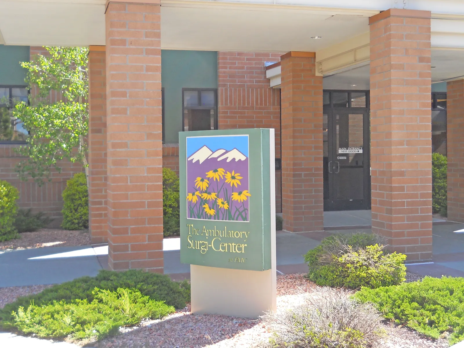 Outdoor sign for The Ambulatory Surgi-Center with flowers and mountain logo in front of brick building entrance.