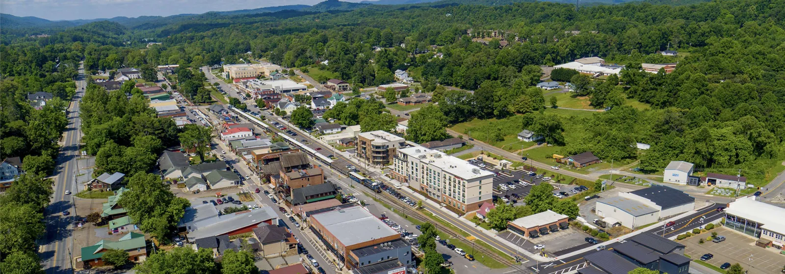 Aerial view of a small town with commercial buildings, streets, and green forested hills in the background.