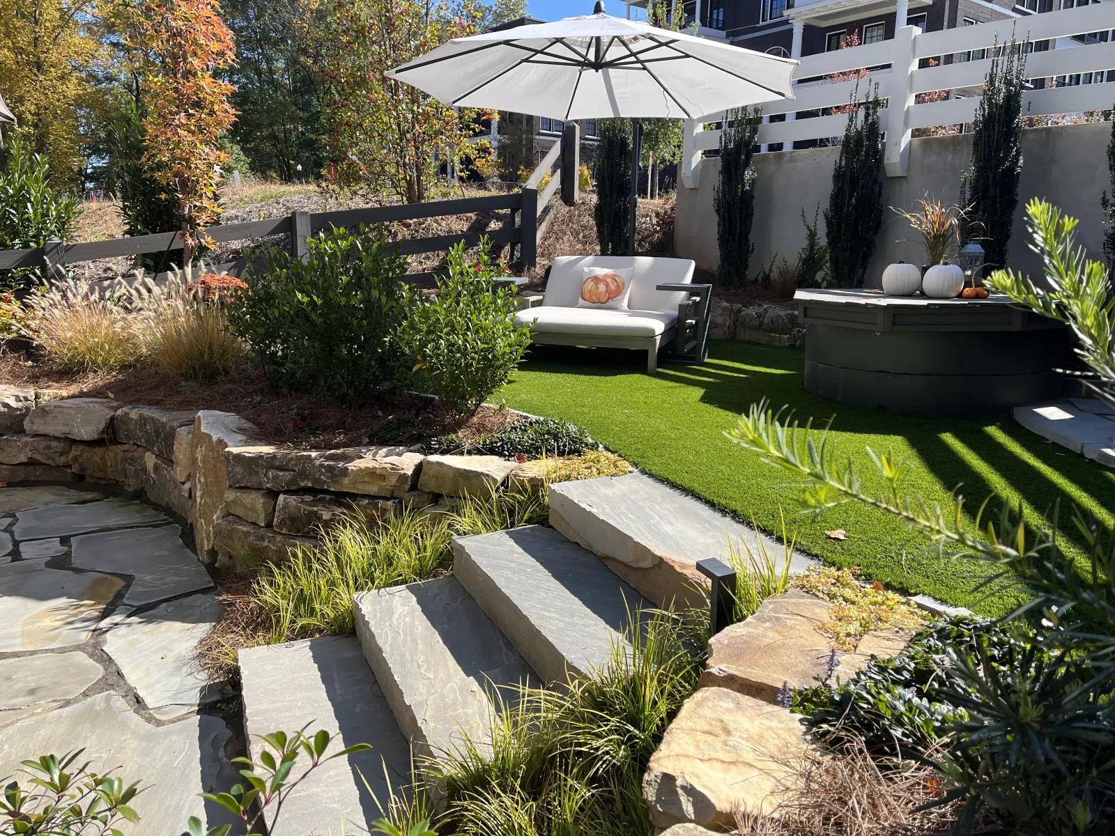 Cozy backyard patio with stone steps, white umbrella, cushioned seating, green grass, and autumn plants.