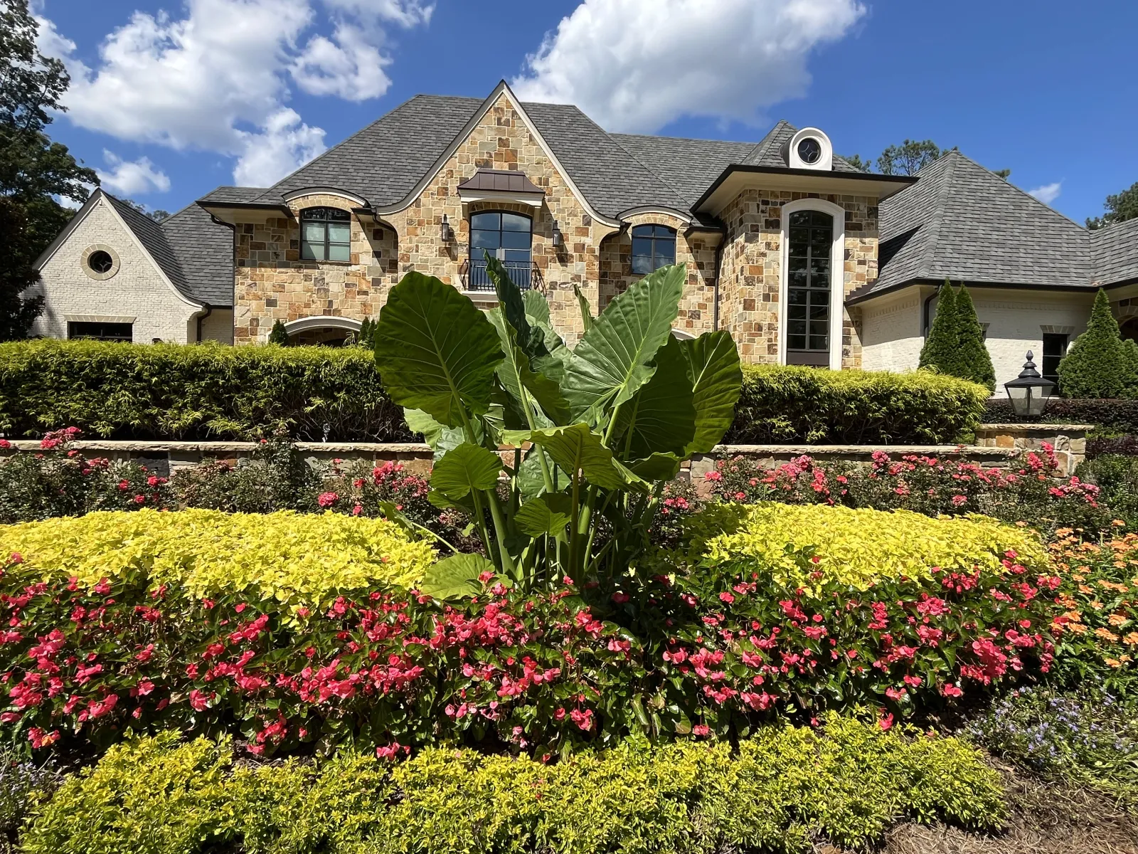 Elegant stone house with manicured garden featuring bright green plants and vibrant pink flowers under blue sky.