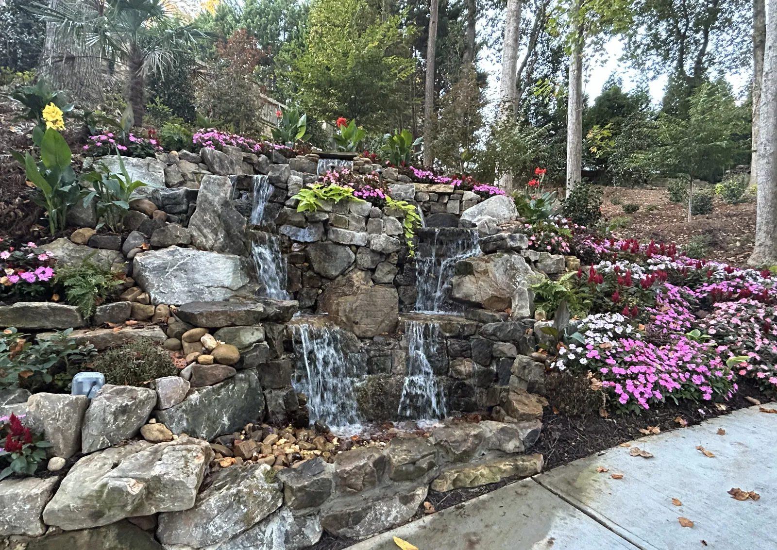 Tiered stone waterfall surrounded by pink and white flowers alongside a wooded garden path on a fall day.