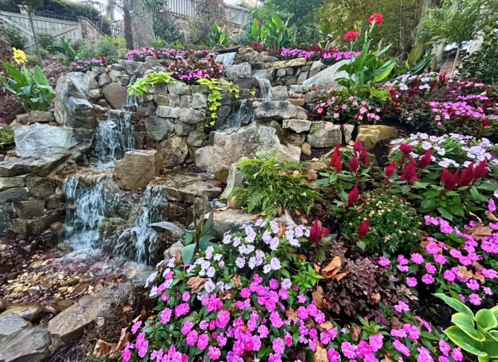 Rock waterfall surrounded by vibrant pink, purple, and red flowers in a lush garden setting.