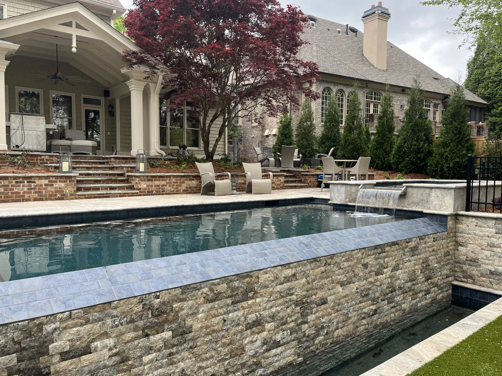 Modern backyard pool with stone walls, waterfall feature, patio seating, and a covered porch beside a large house.