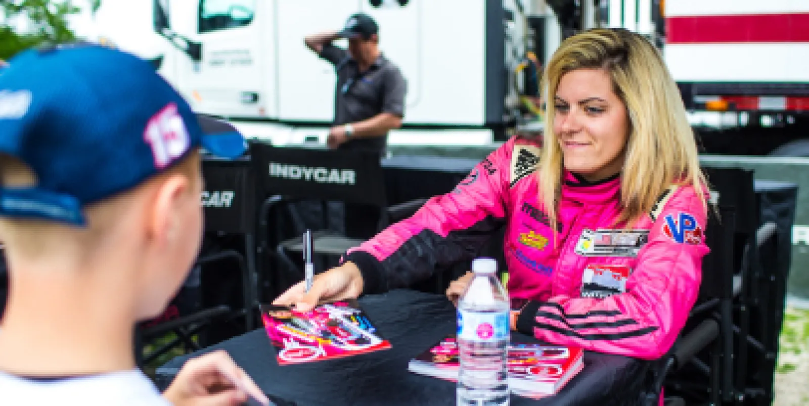 Female race car driver in pink suit signing autographs for a young fan at an IndyCar event.