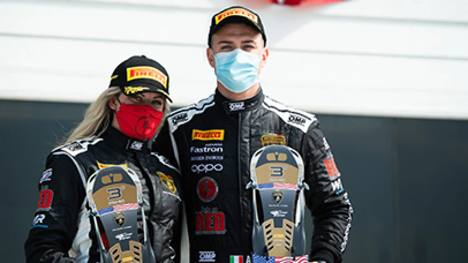 Two masked race car drivers in black suits holding third-place trophies and wearing Pirelli caps on a racetrack.