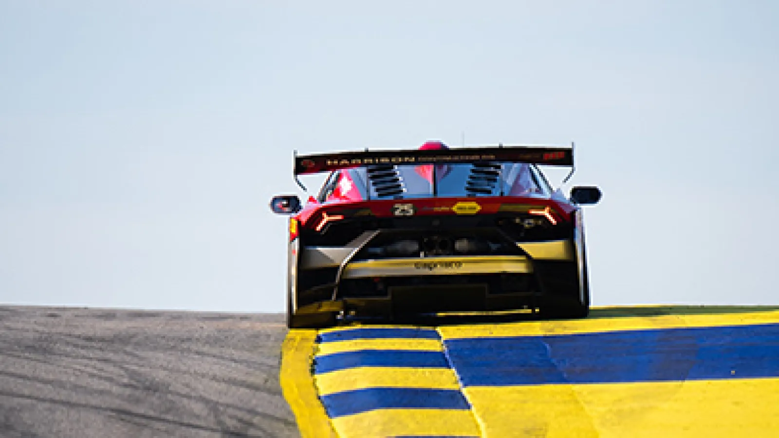 Rear view of a Lamborghini race car speeding on a race track with yellow and blue curbs under a clear sky.
