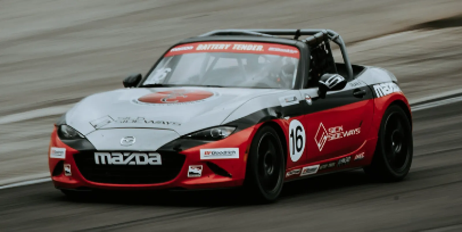 Red, white, and black Mazda race car number 16 speeding on a racetrack with sponsor logos visible