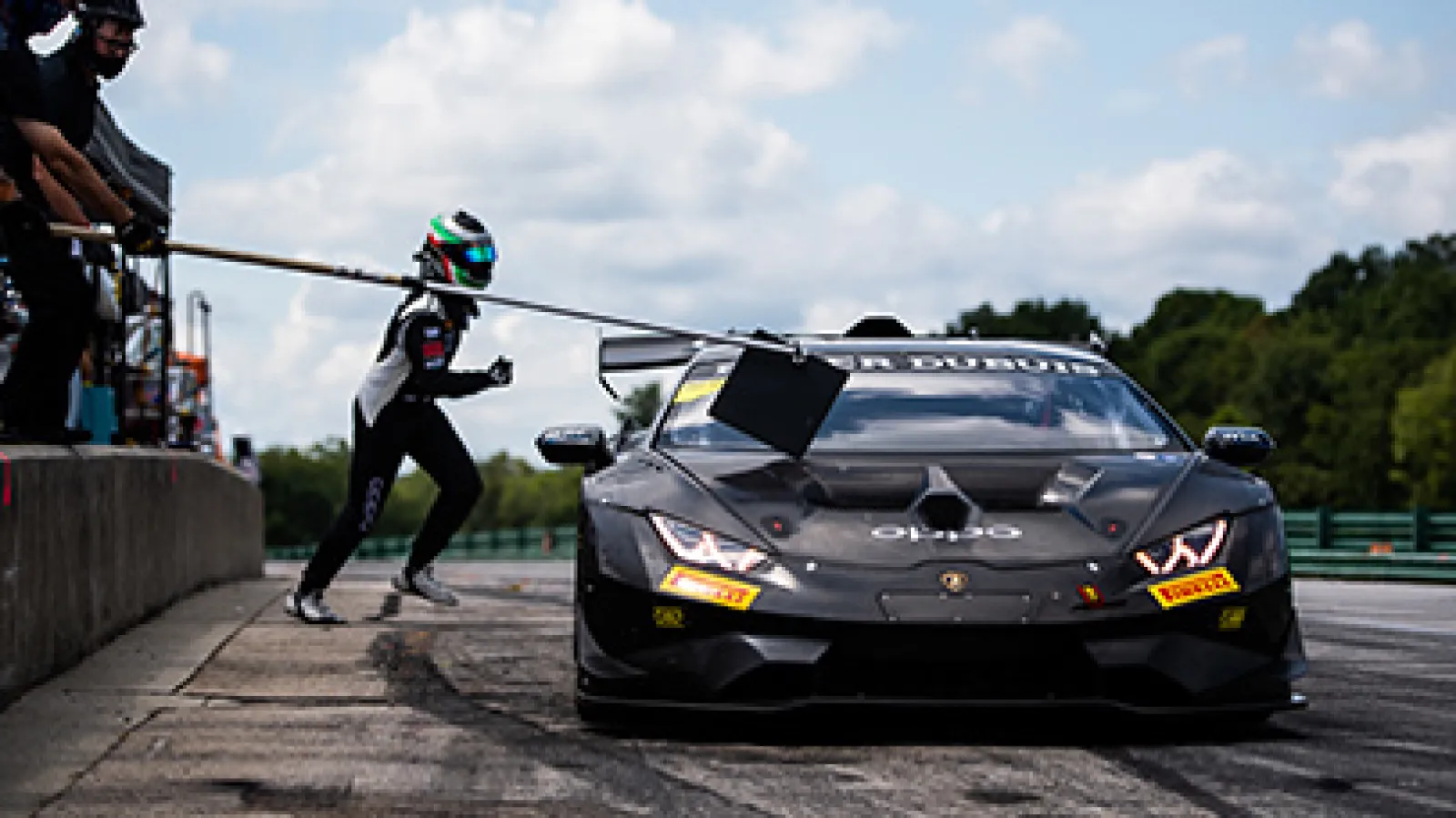 Race car driver exits Lamborghini pit stop during motorsport event under partly cloudy sky.