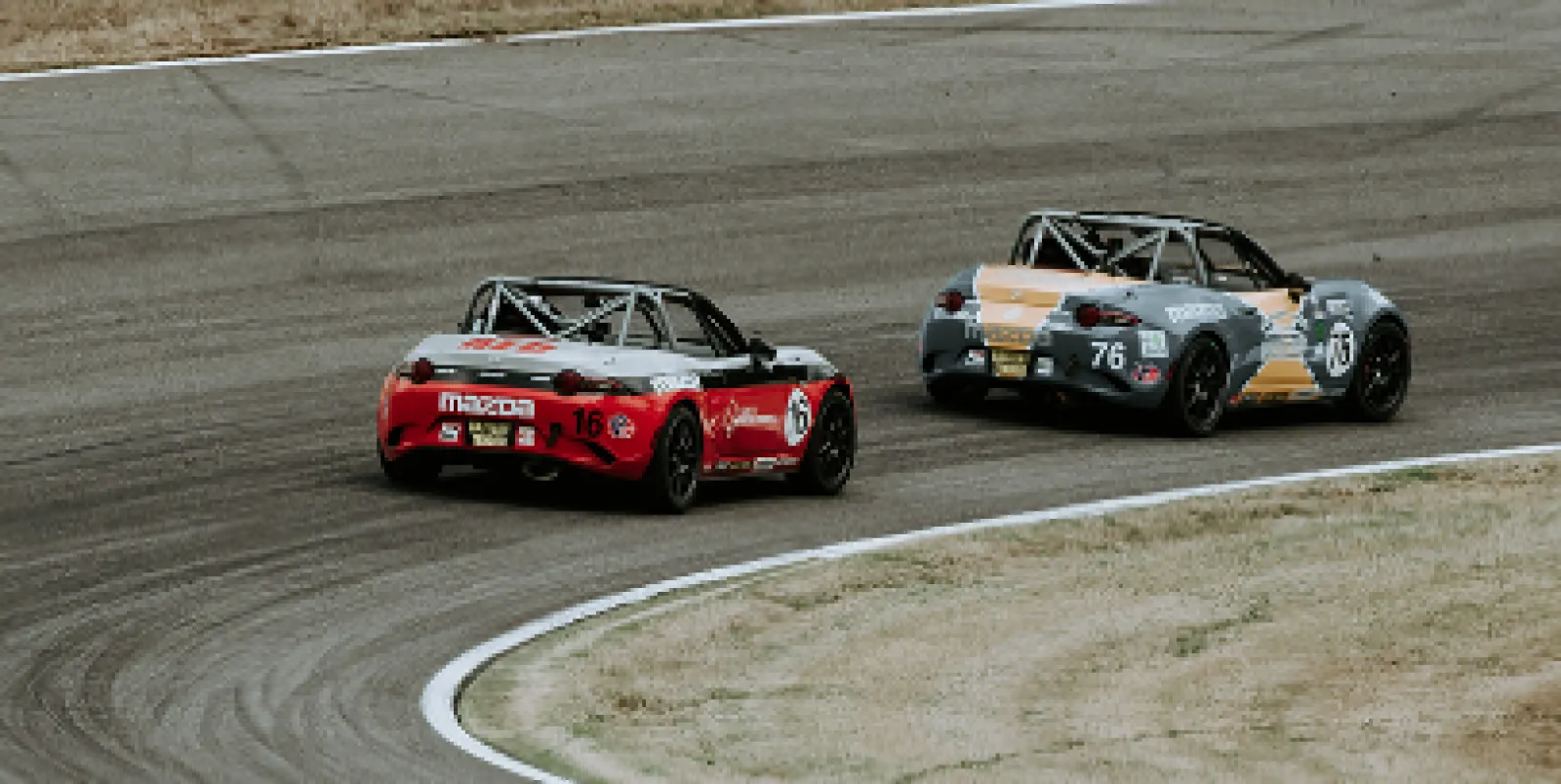 Two Mazda MX-5 race cars, red and gray, competing on a curved asphalt race track in a motorsport event.