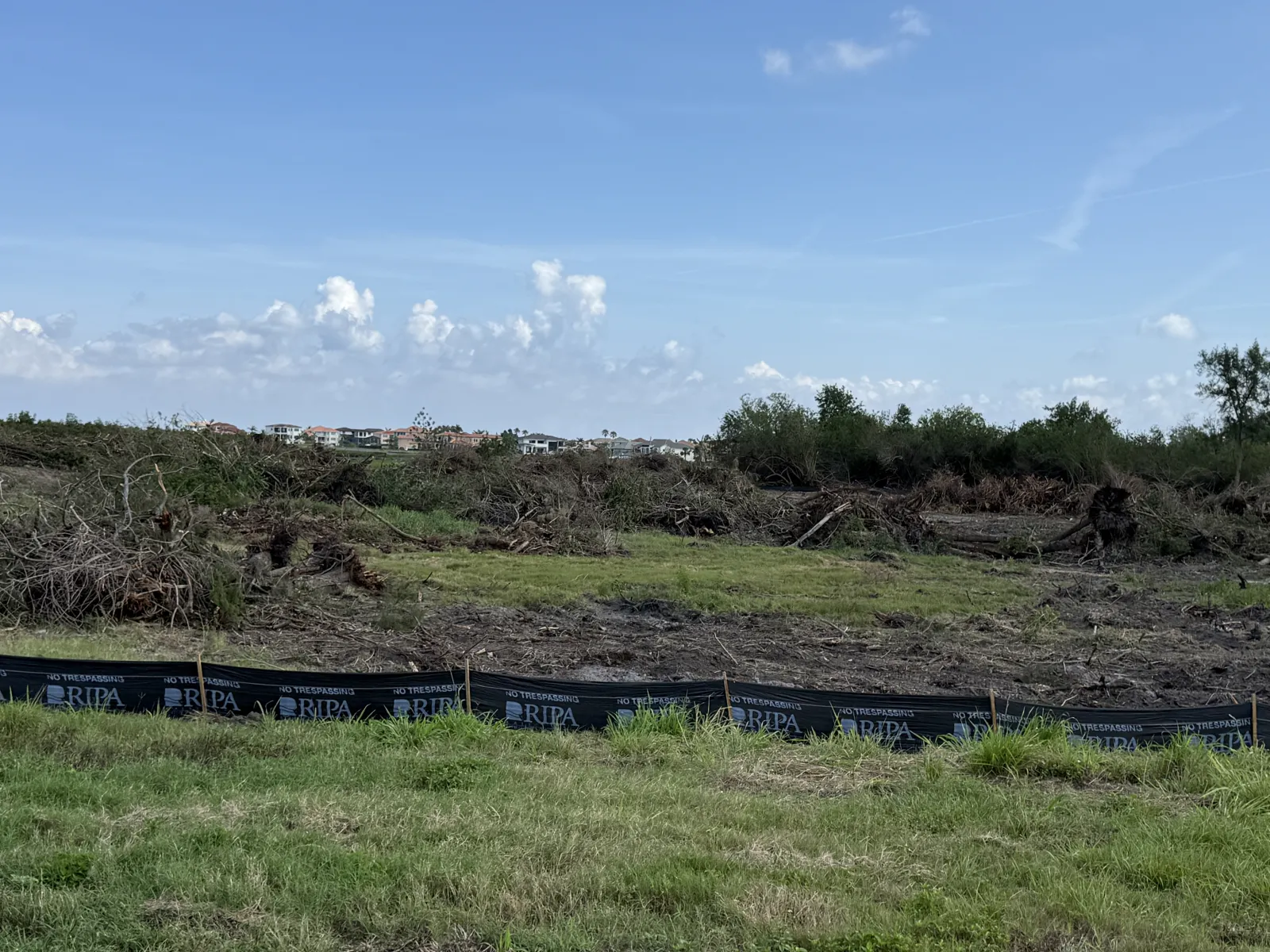 Cleared land with tree debris, grass, and temporary fencing under a partly cloudy blue sky.
