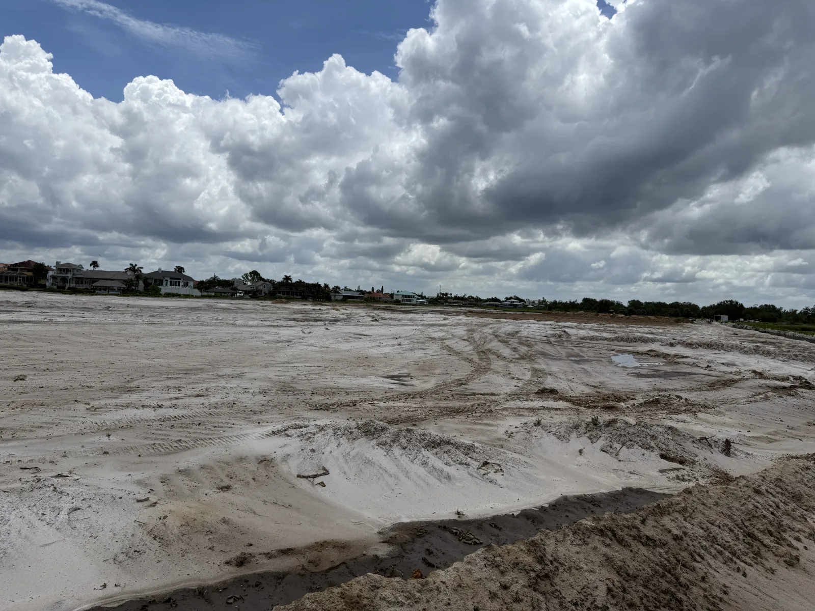 Empty sandy construction site under a cloudy sky near residential houses with visible tire tracks.