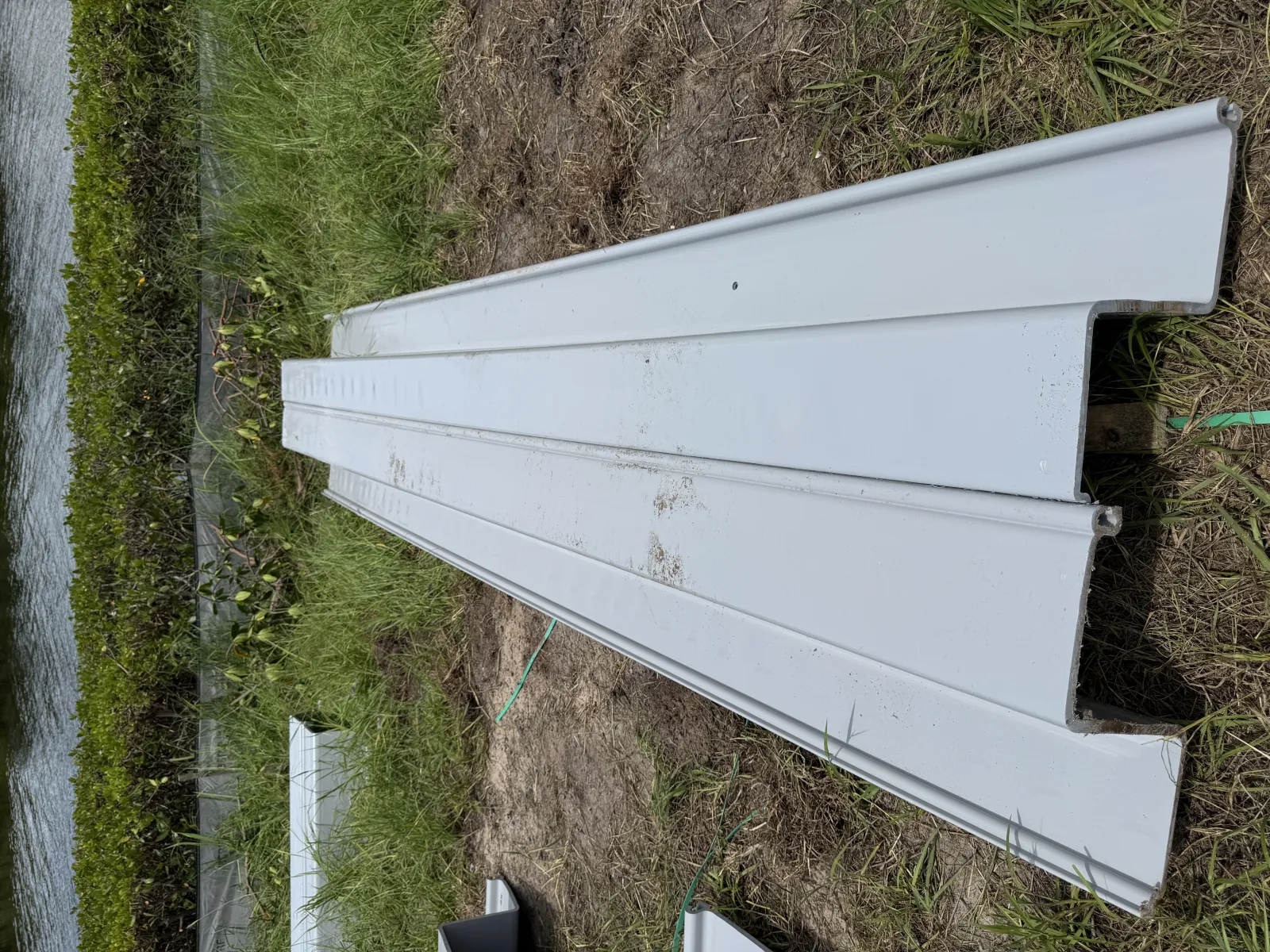 White metal roofing panels stacked on grassy ground near a water body and plants.