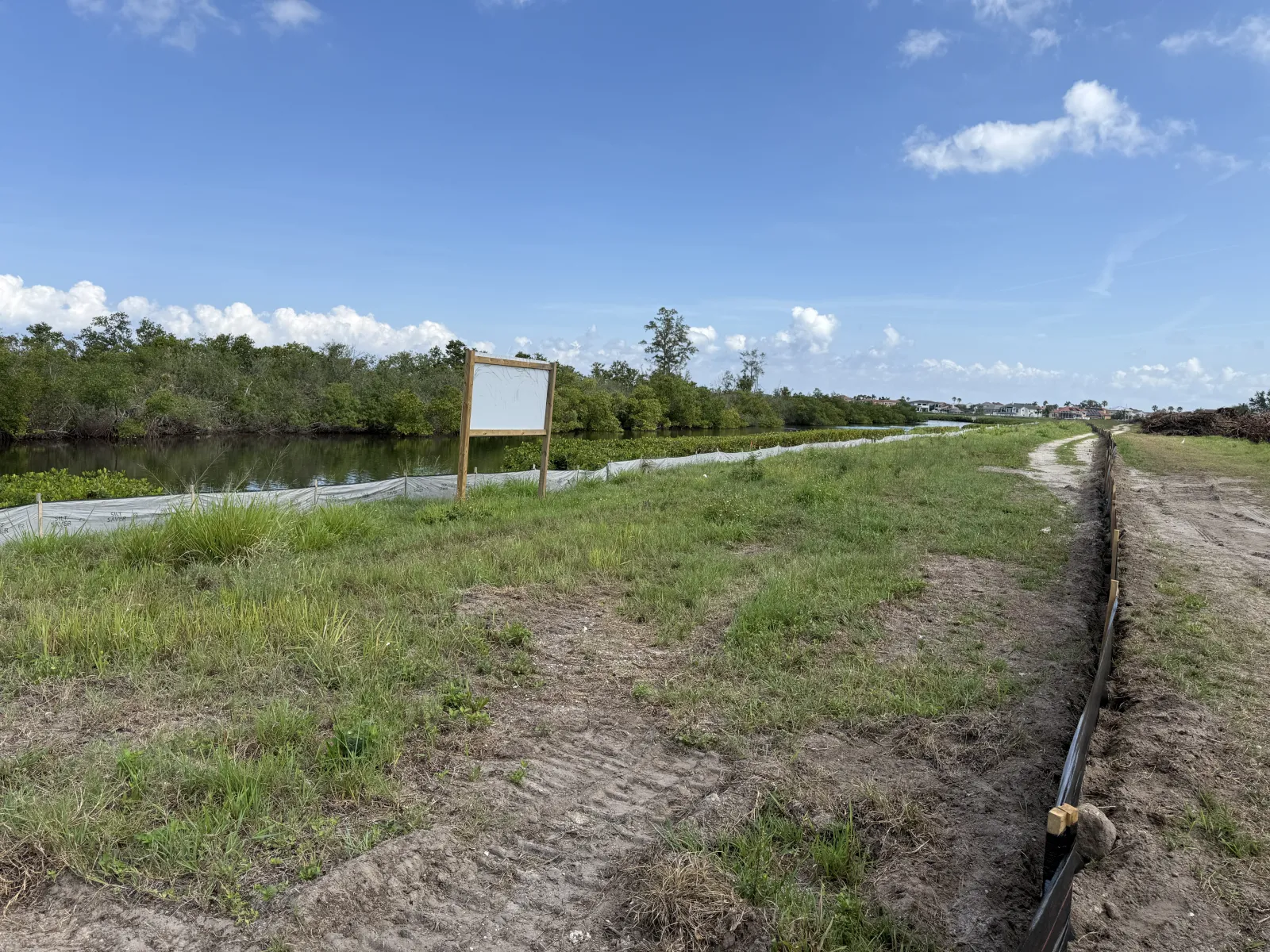 Grassy landscape with a small water channel, a blank sign, and a dirt path under blue sky with clouds