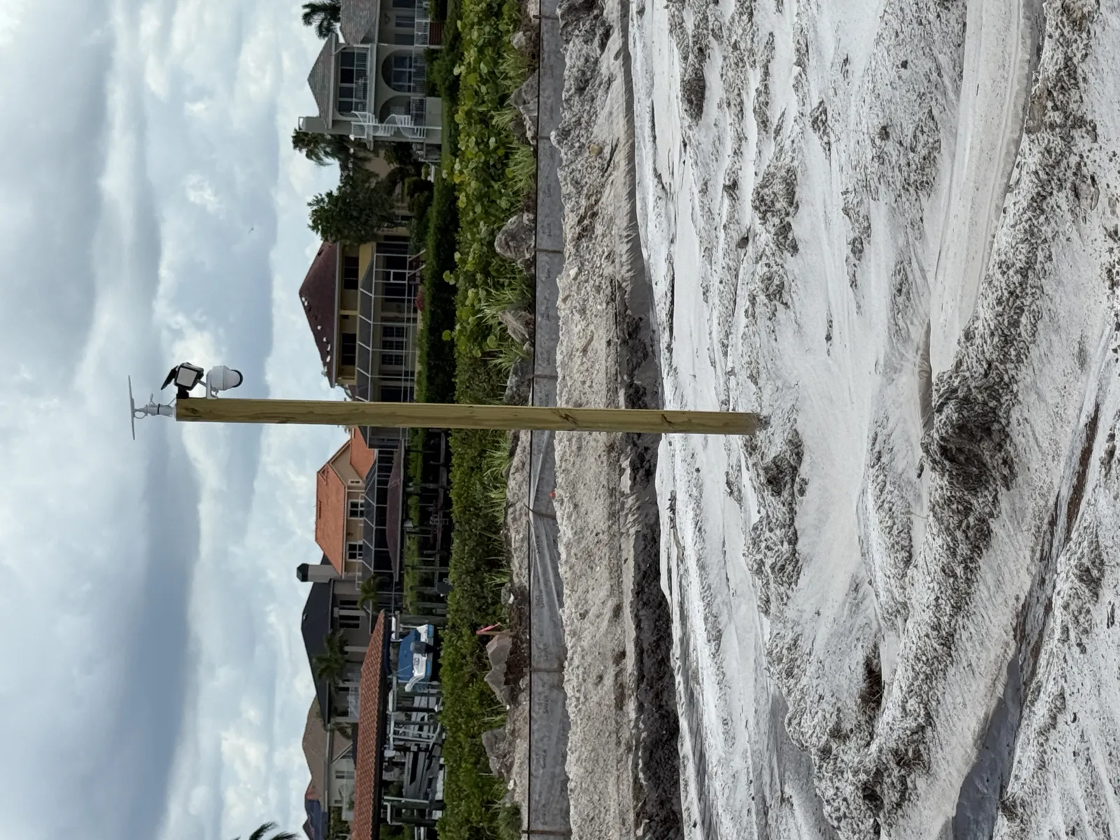 Wooden pole with mounted devices on sandy ground near residential houses and cloudy sky