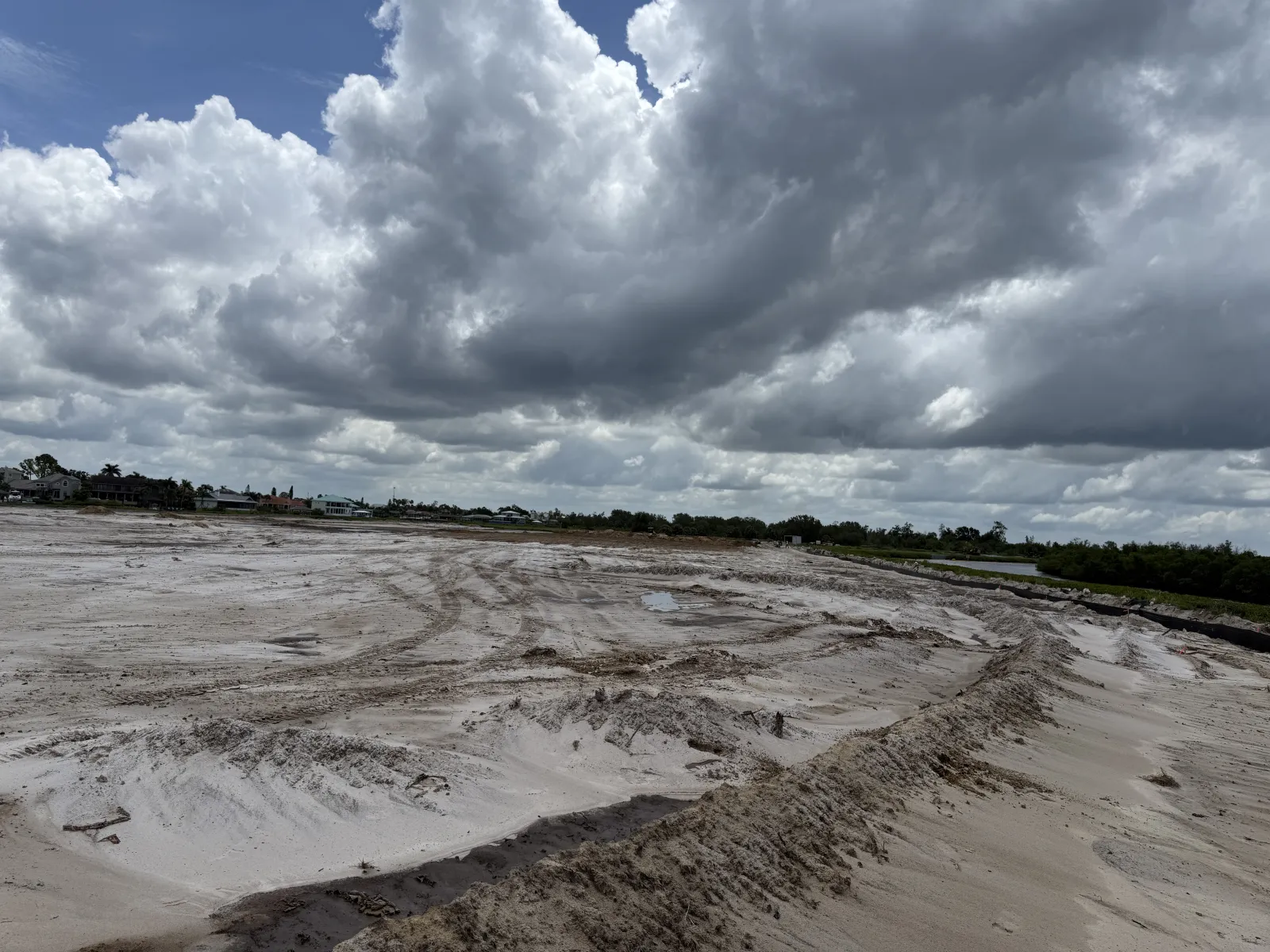 Wide sandy construction site under a dramatic cloudy sky with distant houses and greenery on horizon.