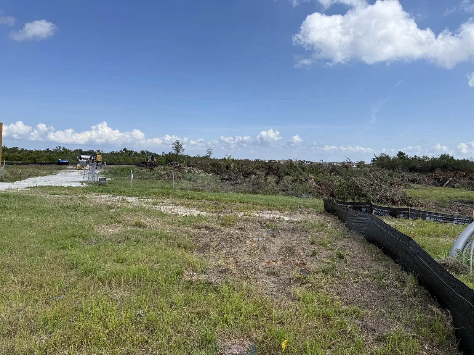 Cleared construction site with grassy foreground, dirt patches, machinery, and blue sky with clouds.