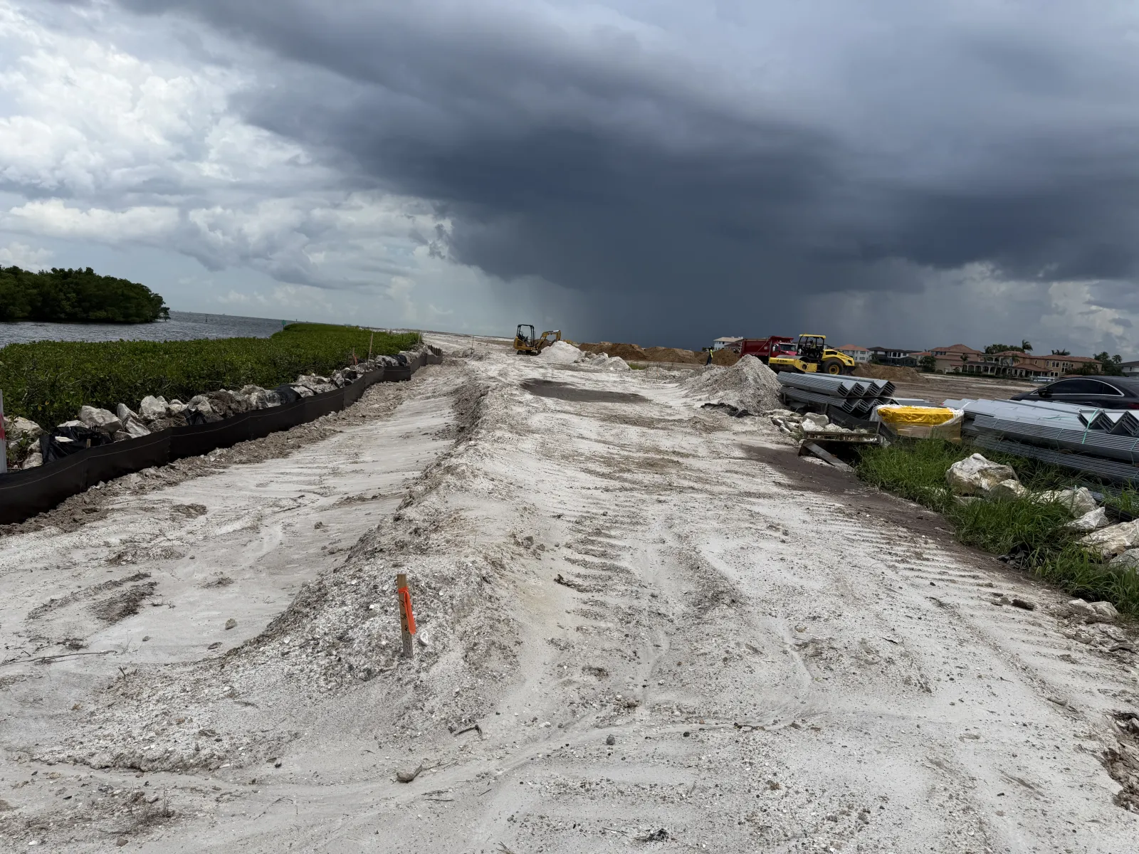 Construction site with dirt road and equipment under dark stormy clouds near waterfront and vegetation.