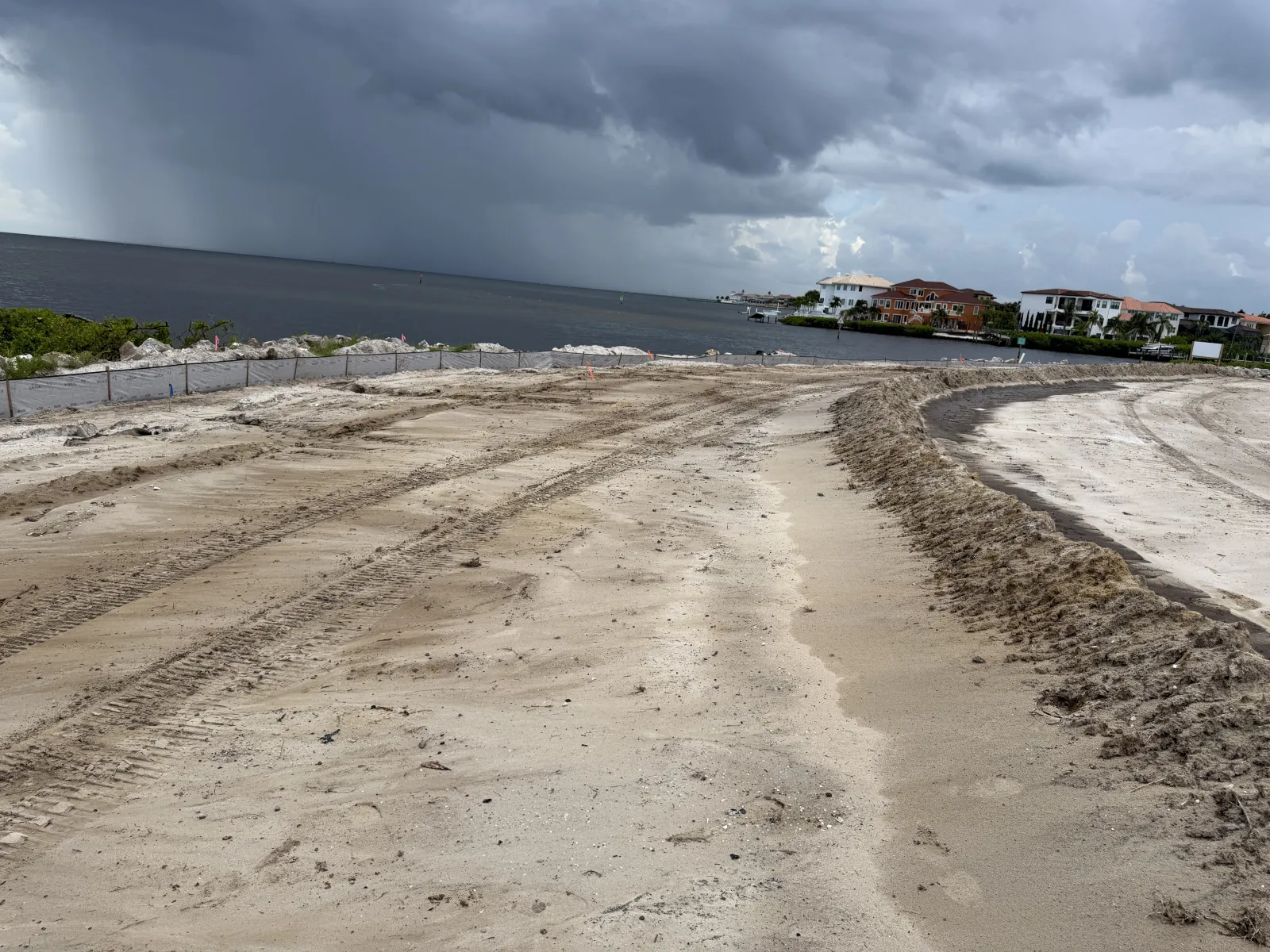 Construction site by the water with sandy terrain and heavy tire tracks under stormy cloudy sky.