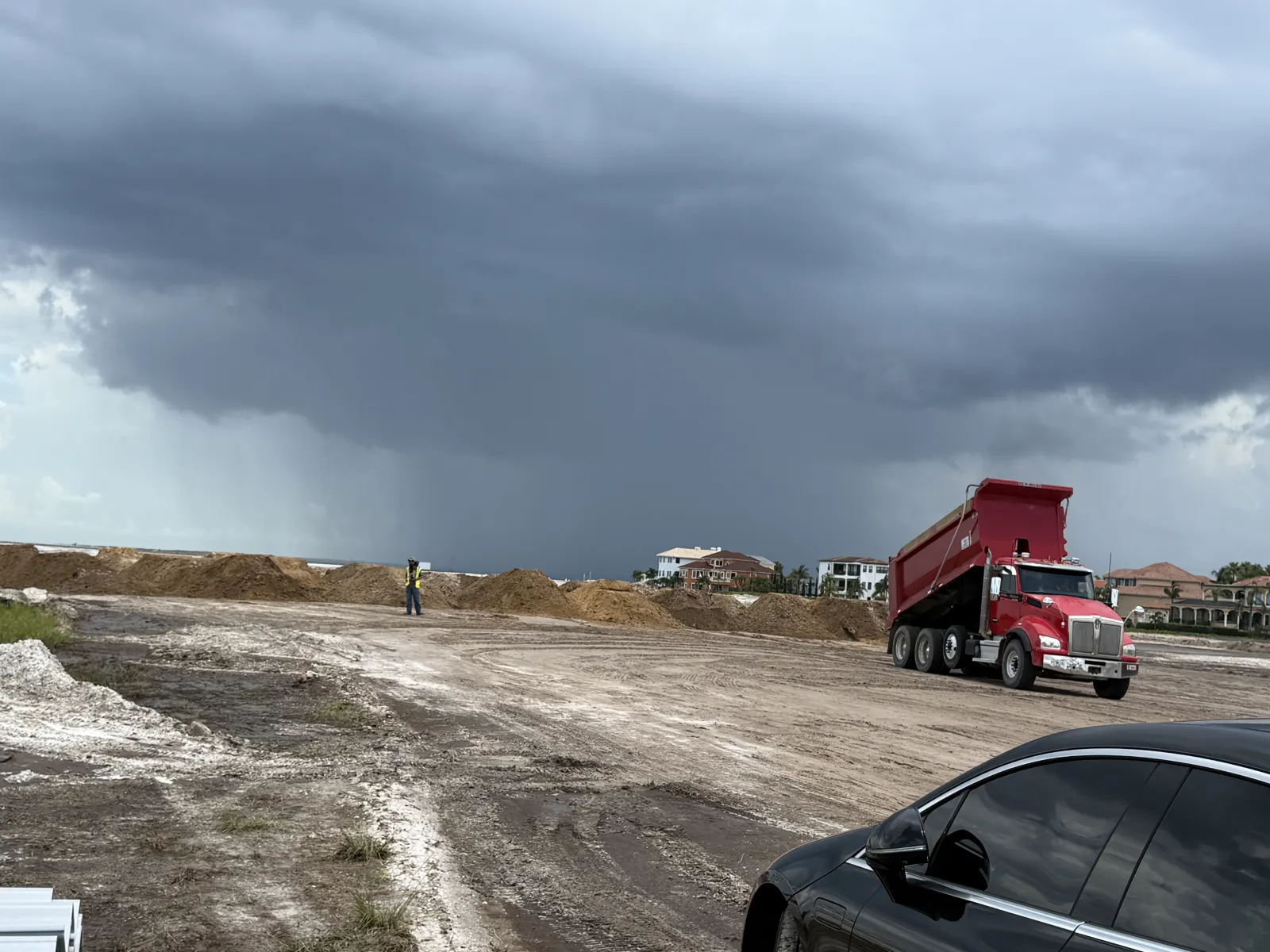 Red dump truck unloading soil on construction site under dark rainy clouds with worker and houses in background.