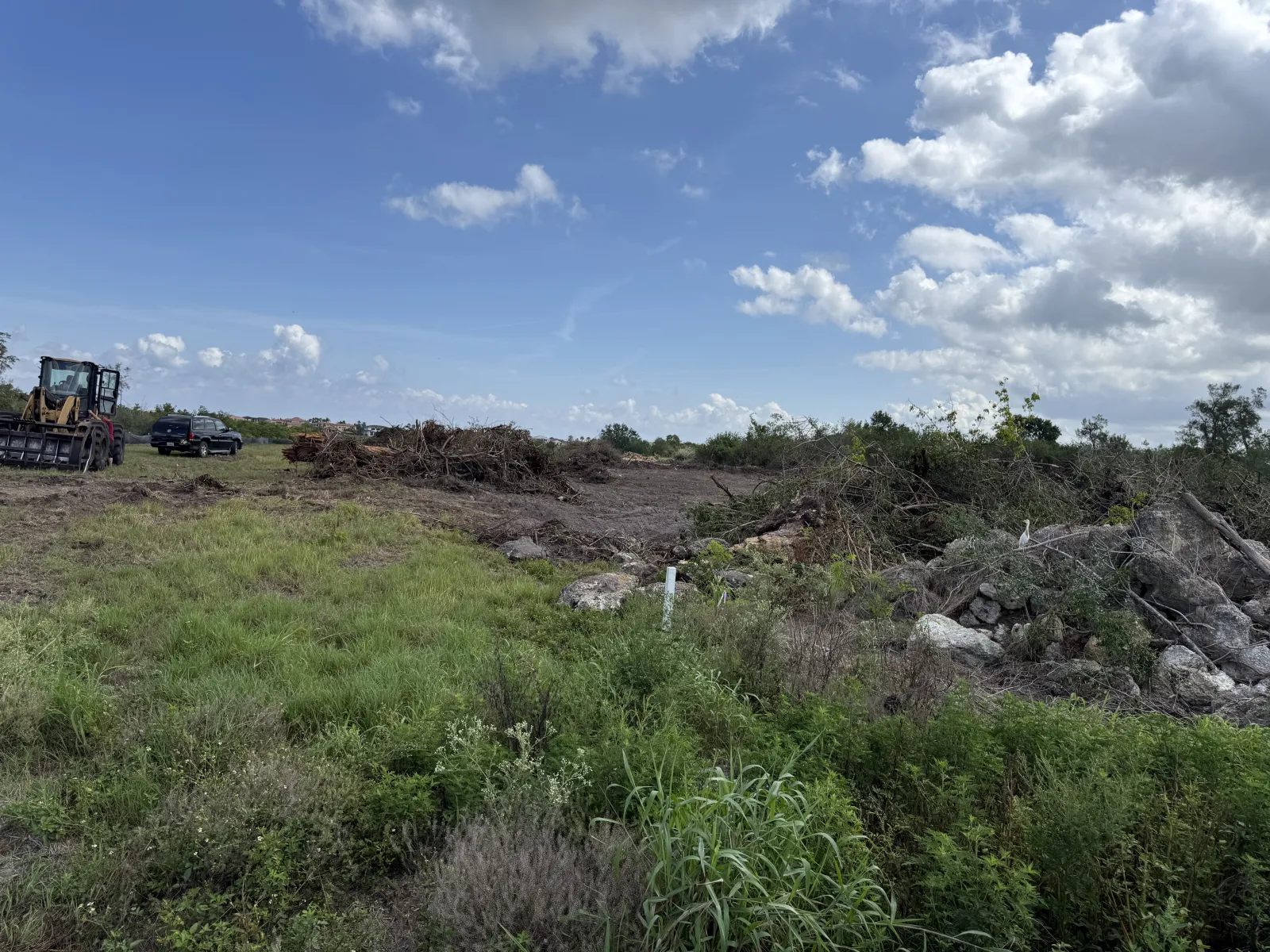 Cleared land with piles of debris and construction equipment under a partly cloudy blue sky.