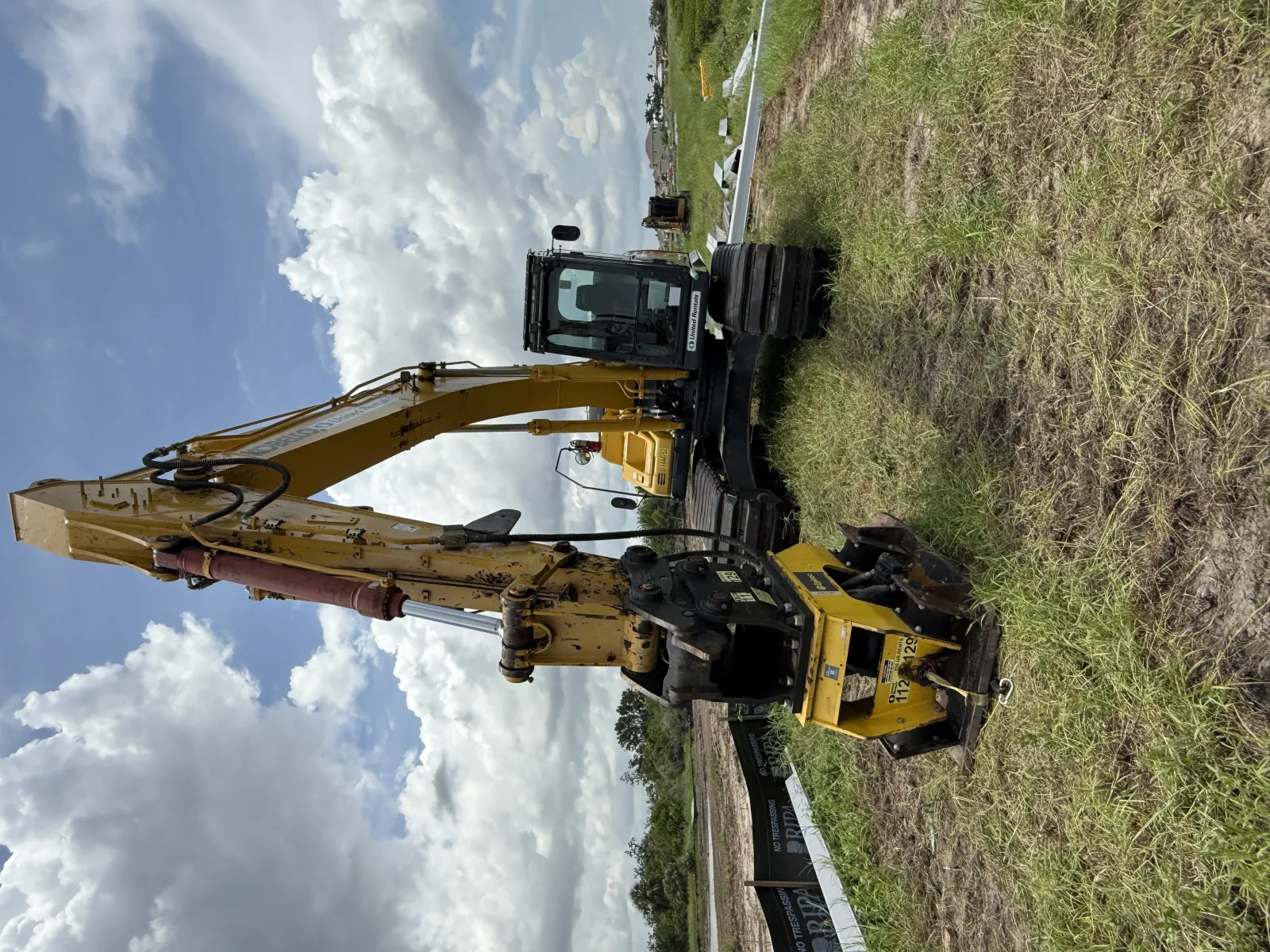 Yellow excavator with hydraulic arm and metal attachments parked on grassy field under cloudy sky.