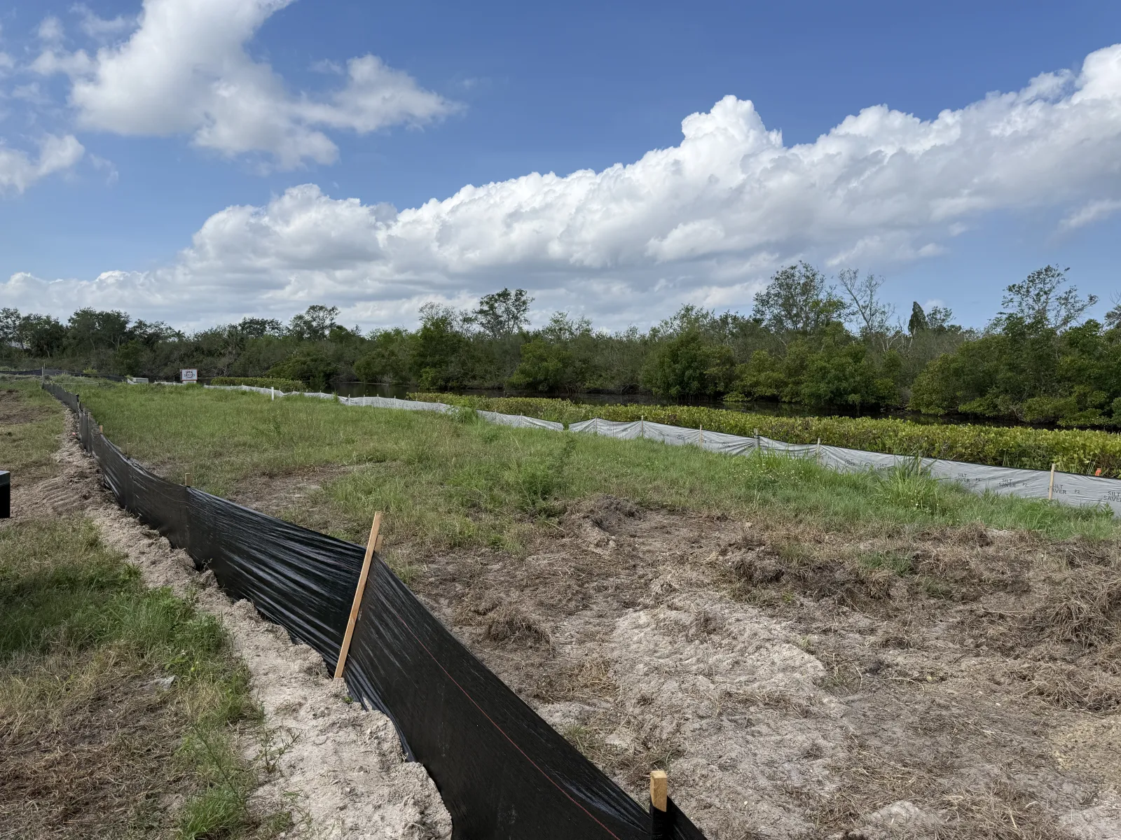 Construction site barrier fencing separates disturbed soil from grassy area under a blue sky with clouds.