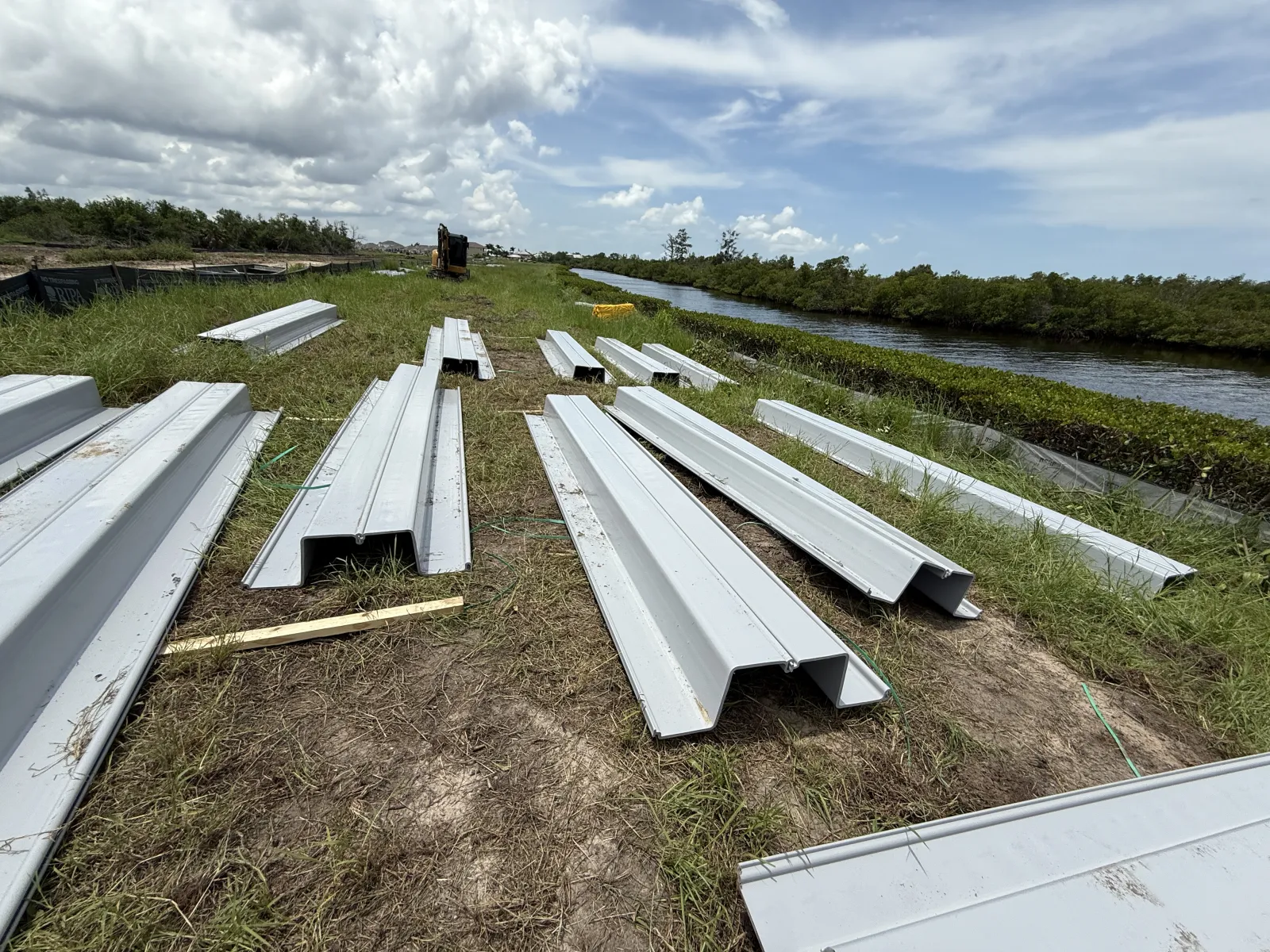 Metal sheet piles laid out on grass near a canal under a cloudy sky at a construction site.