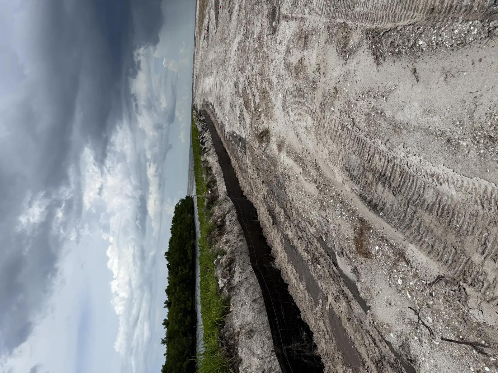 Construction site with excavated trench, sandy ground, and cloudy sky over a green landscape.